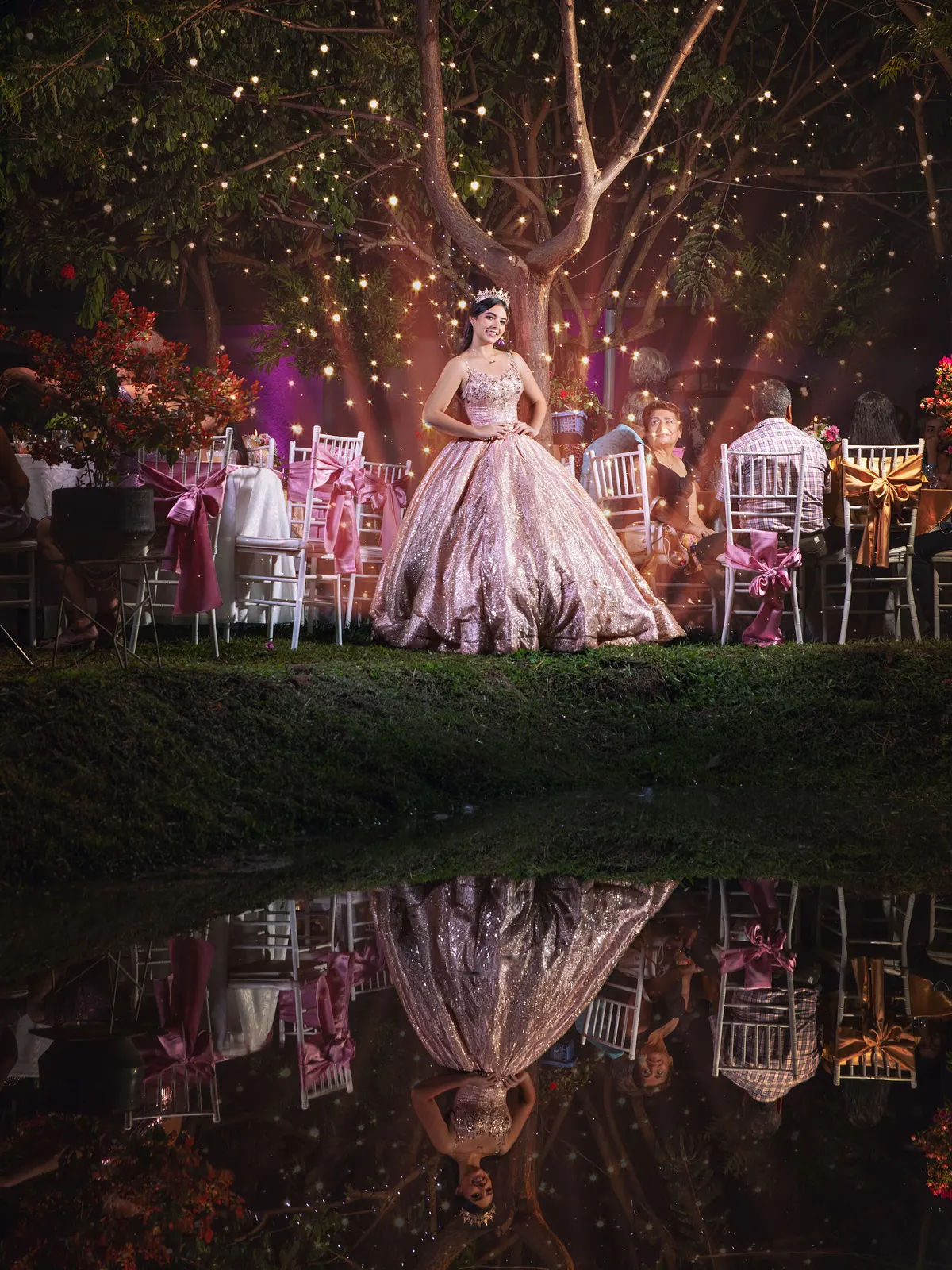 Quinceañera con vestido rosa reflejada en el agua bajo luces de un árbol