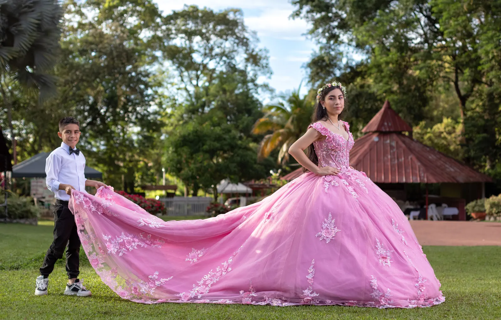Quinceañera con vestido rosa de cola larga junto a un niño en un jardín