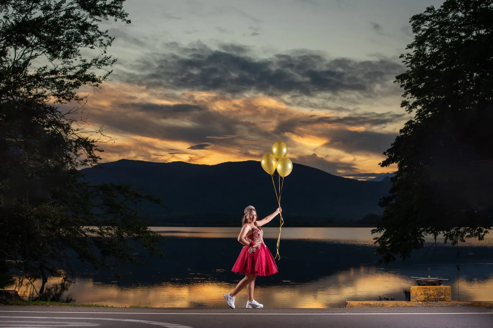 Quinceañera con vestido rojo y globos dorados junto al lago al atardecer